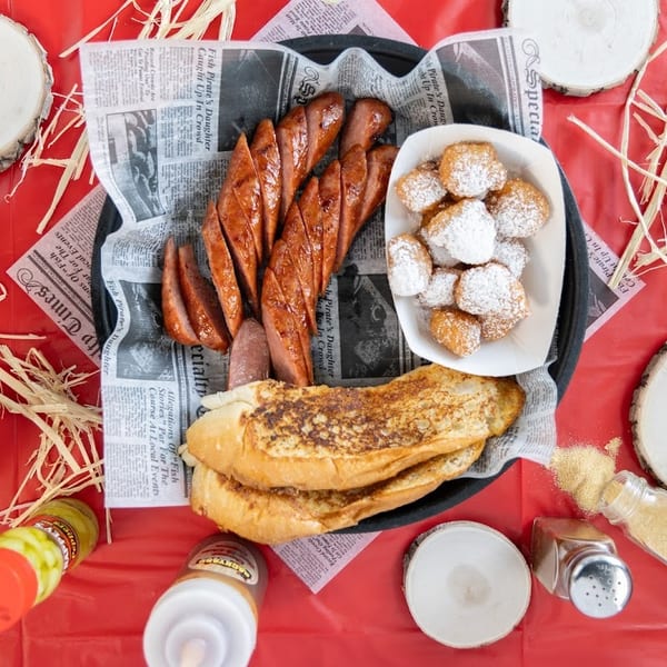Overhead view of grilled sausage, beignets, and bread on a newspaper-lined tray with red background.